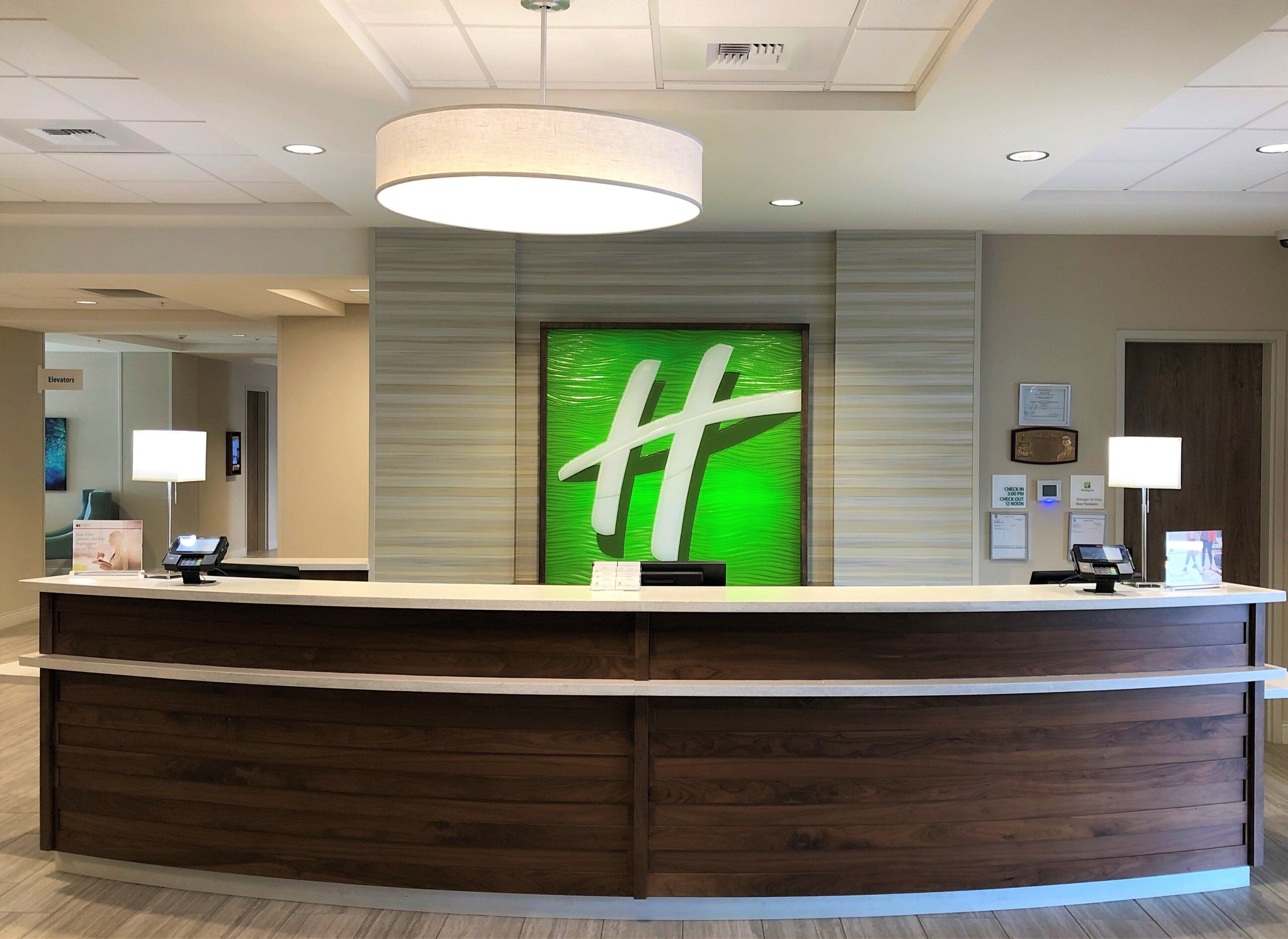 A hotel reception desk with a modern design, a green sign with a white 'H' logo, and a digital check-in kiosk.