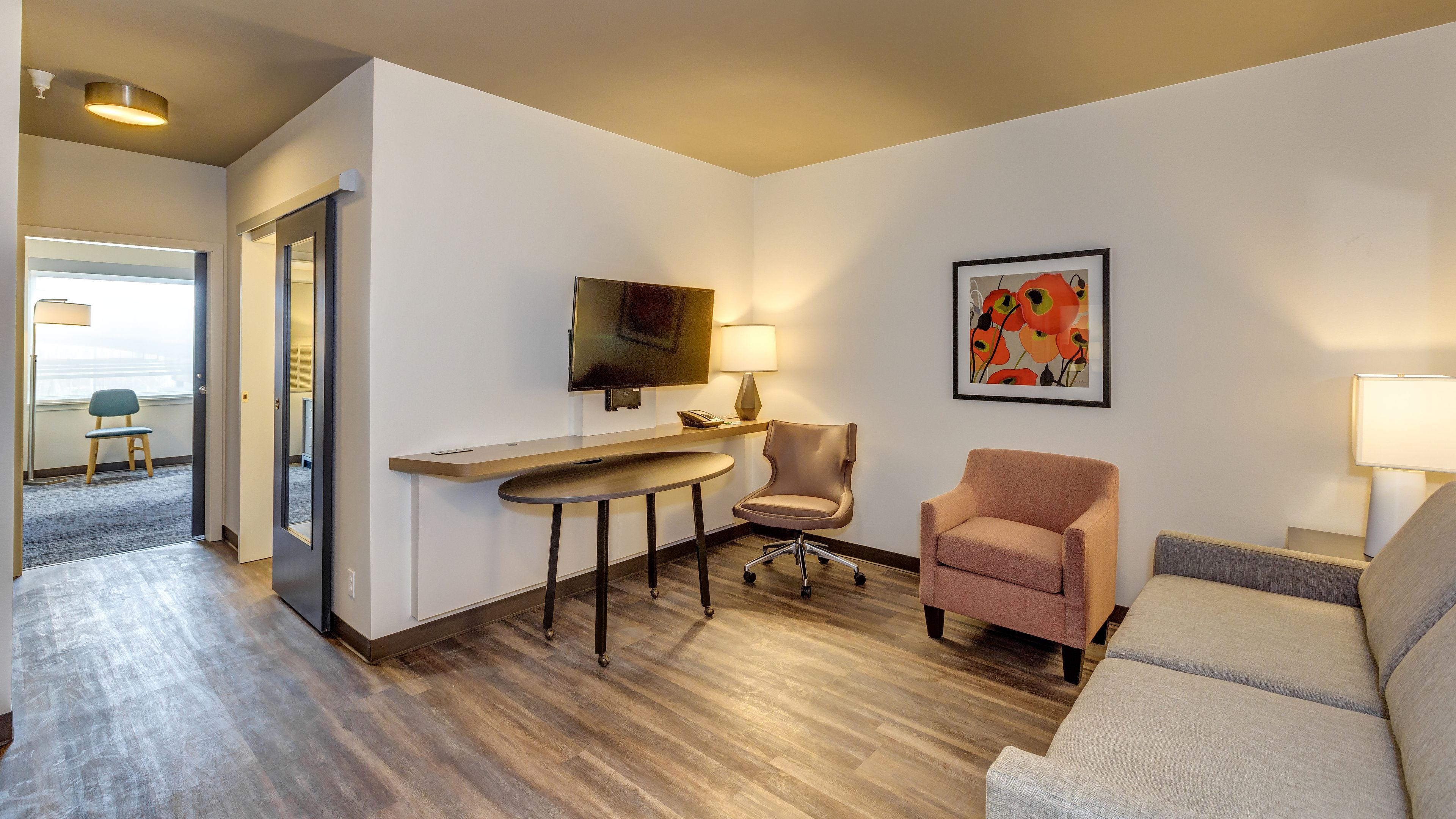 Habitación de hotel con piso de madera, un escritorio con silla, un sofá, un televisor montado en la pared y una obra de arte enmarcada.