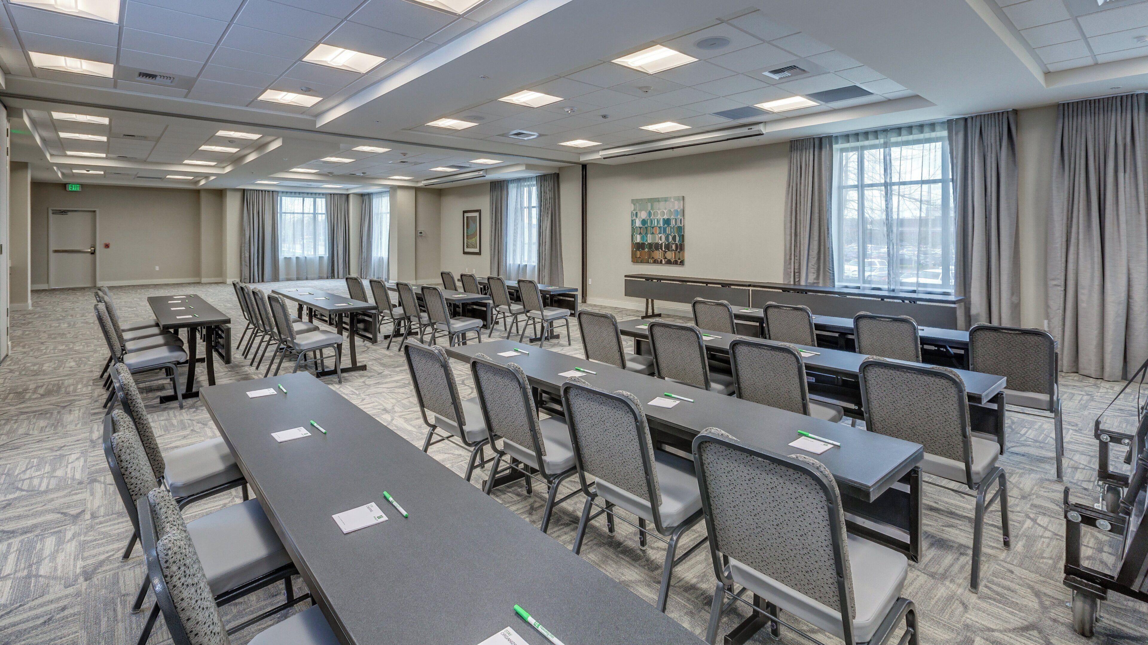 A conference room with multiple tables and chairs, large windows, and a carpeted floor.
