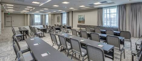 A conference room with multiple tables and chairs, large windows, and a carpeted floor.