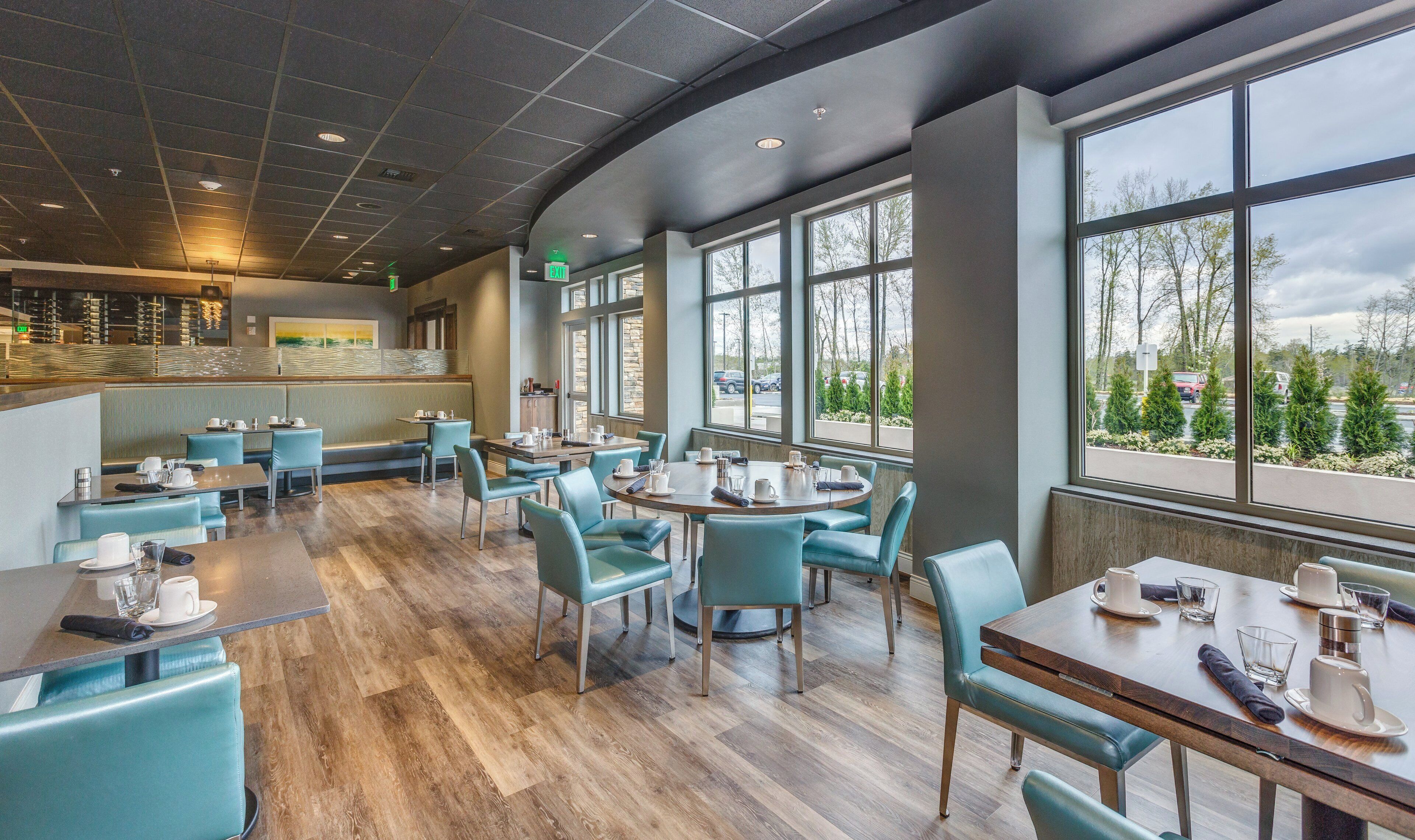 Un espace de restauration moderne avec un plancher en bois, des chaises de couleur turquoise et de grandes fenêtres donnant sur le parking et les arbres.