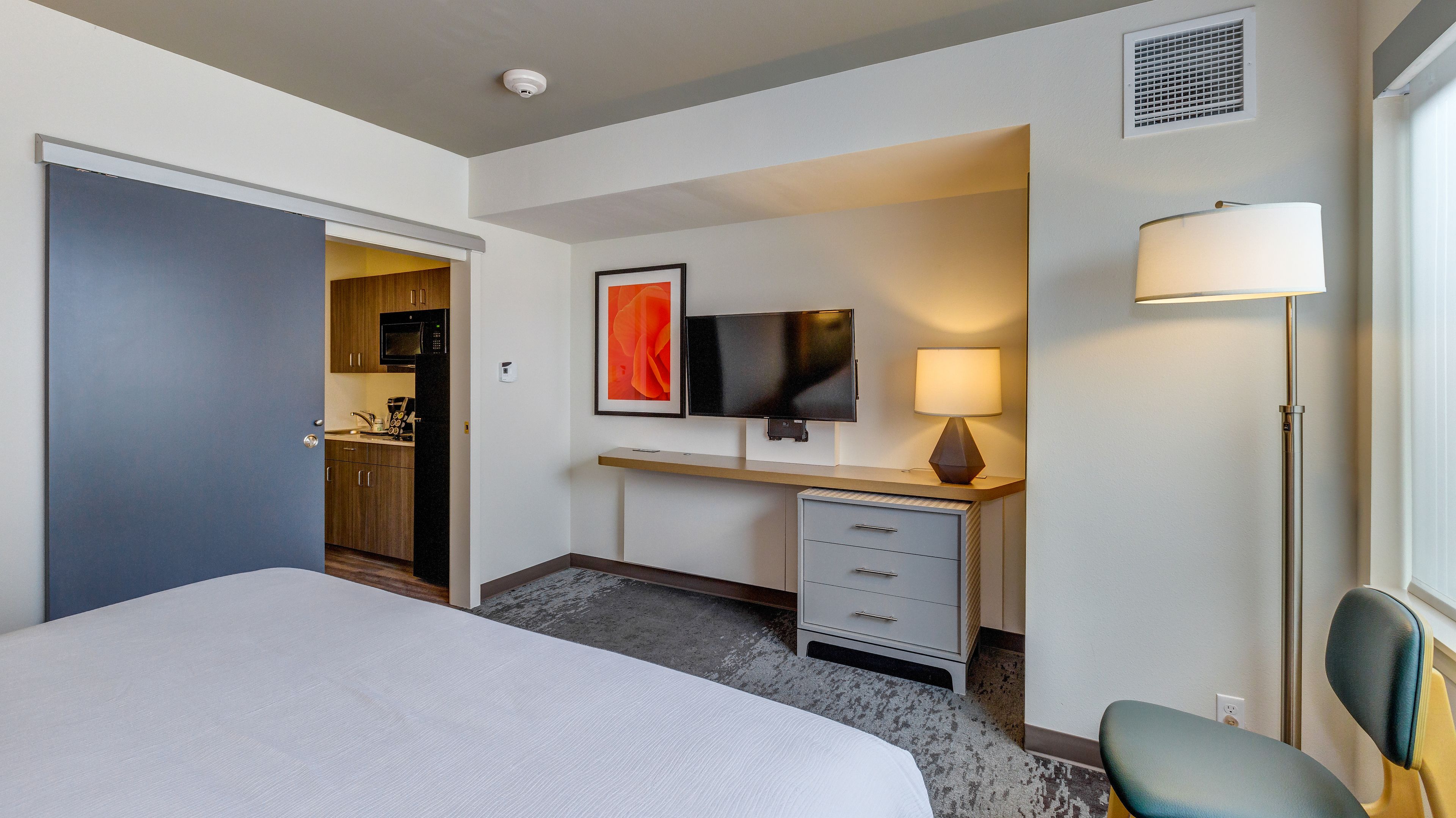 A hotel room with a bed, a desk with a lamp, a television, a minibar, and a door leading to a kitchenette.