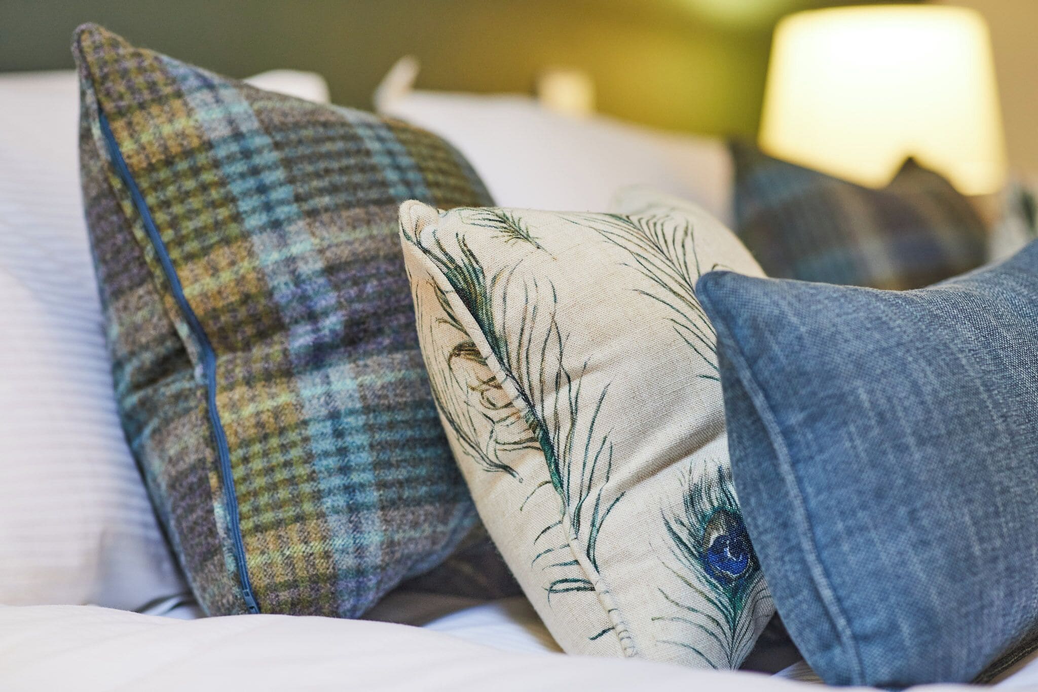 A bed with multiple pillows, including a plaid one and a feathered one, and a pair of jeans.