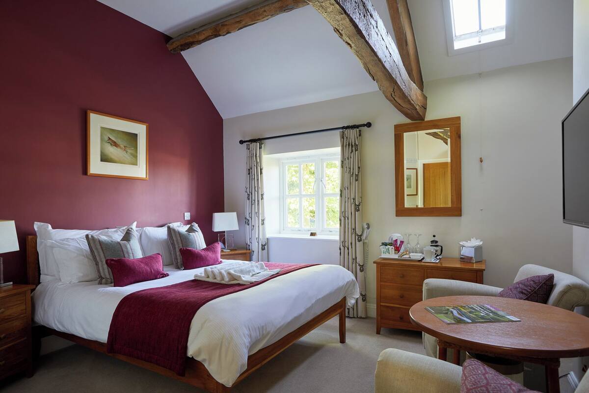 A bedroom with a large bed, a wooden headboard, a bedside table with a lamp, a dresser, a round table with a magazine, and a window with curtains.