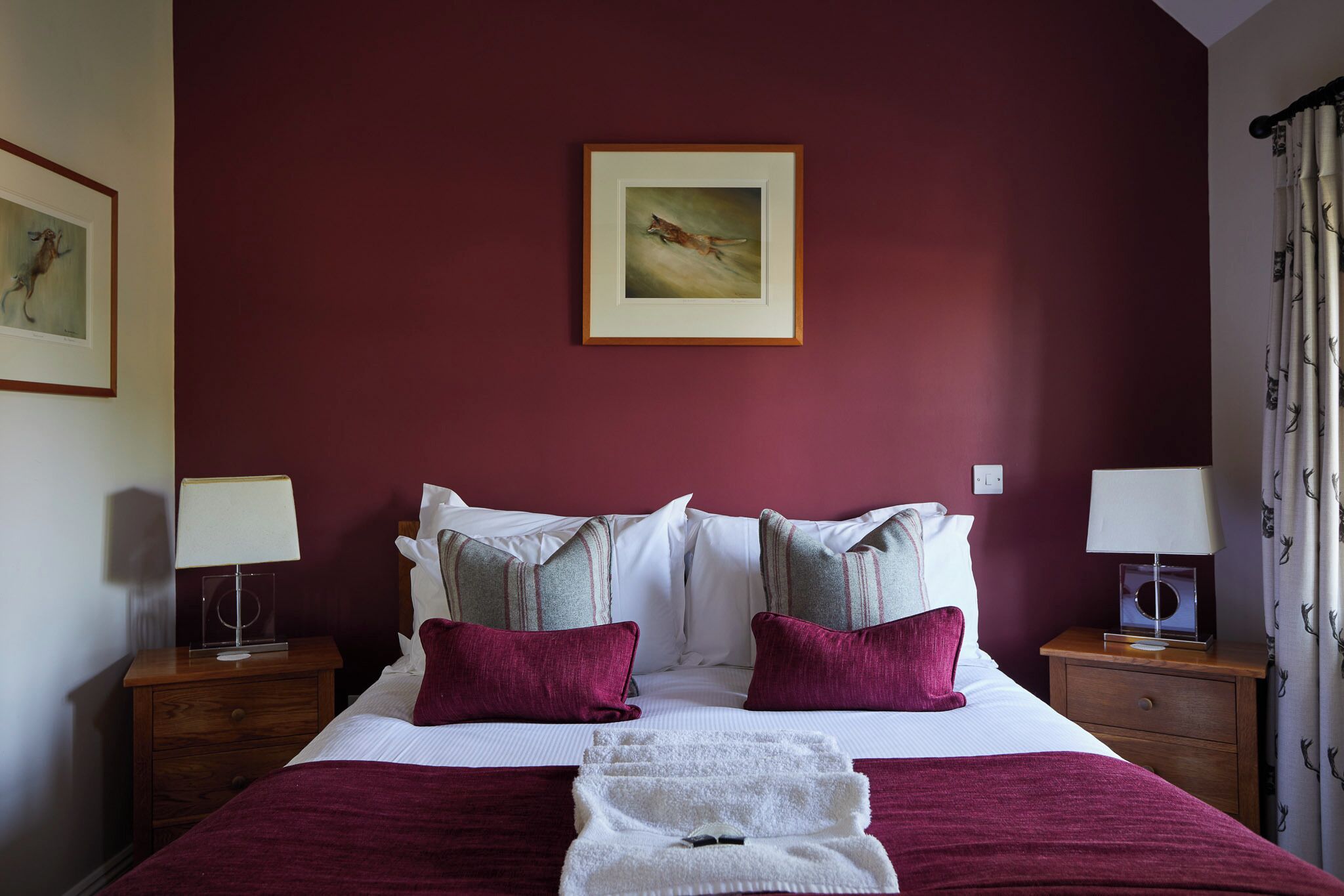 Una cama bien hecha con ropa de cama blanca y color burdeos, dos mesitas de noche con lámparas, un cuadro en la pared y una ventana con cortinas.