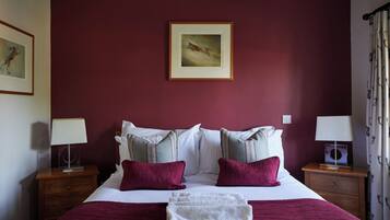 Una cama bien hecha con ropa de cama blanca y color burdeos, dos mesitas de noche con lámparas, un cuadro en la pared y una ventana con cortinas.