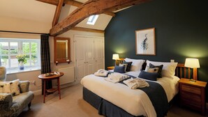 A bedroom with a large bed, two bedside tables with lamps, a wooden ceiling beam, a window with curtains, and a chair. - The Old Coach House (Ripon)
