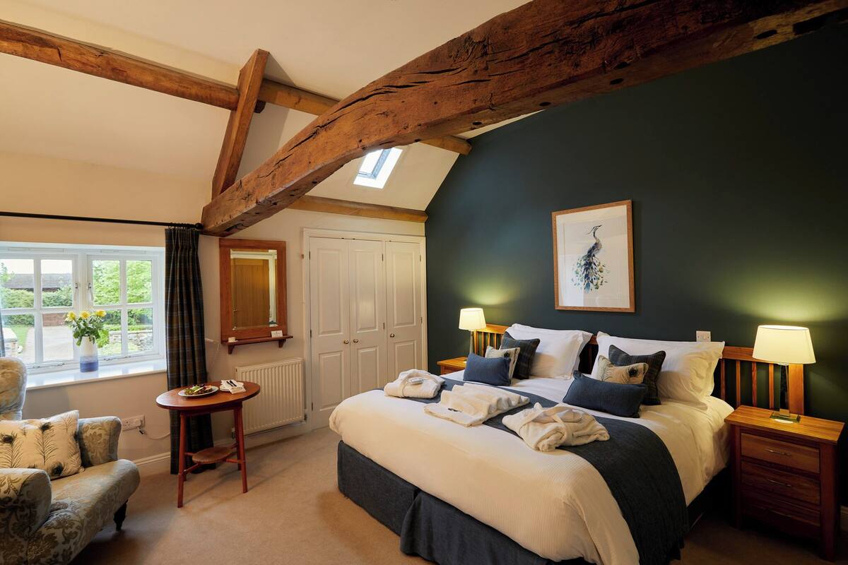 A bedroom with a large bed, two bedside tables with lamps, a wooden ceiling beam, a window with curtains, and a chair.
