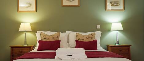 Una cama bien hecha con decoración en blanco y rojo, flanqueada por dos mesitas de noche con lámparas, y una obra de arte en la pared.