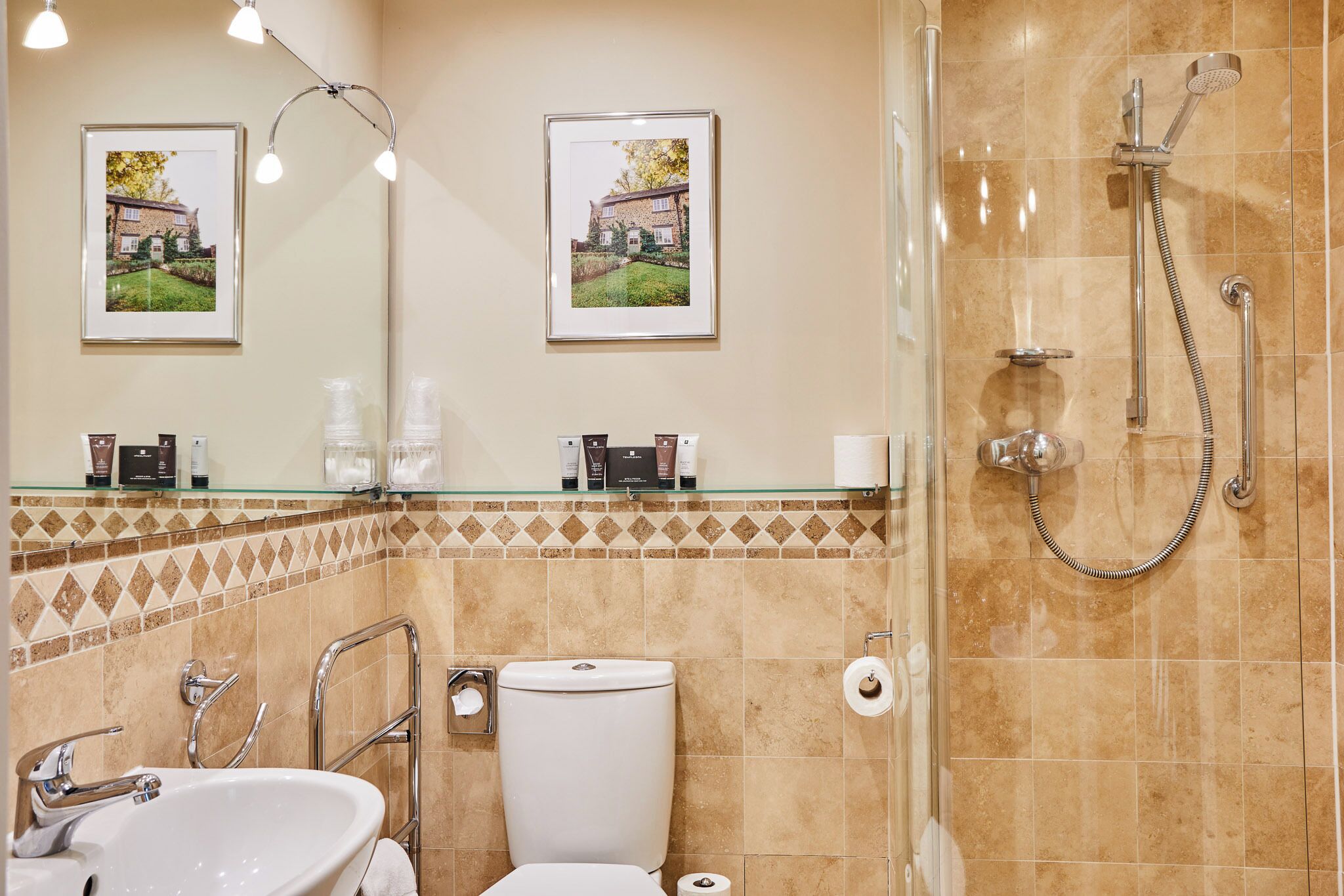 A bathroom with a glass shower enclosure, a white toilet, a sink, and two framed pictures on the wall.