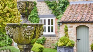 A stone fountain with water spouting from its top, set against a stone building with a window and door. - The Old Coach House (Ripon)