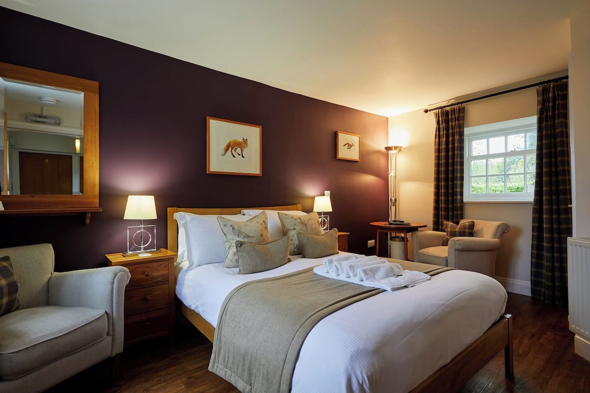 A well-lit hotel room with a large bed, a bedside table with a lamp, a chair, a small table, and a window with curtains.