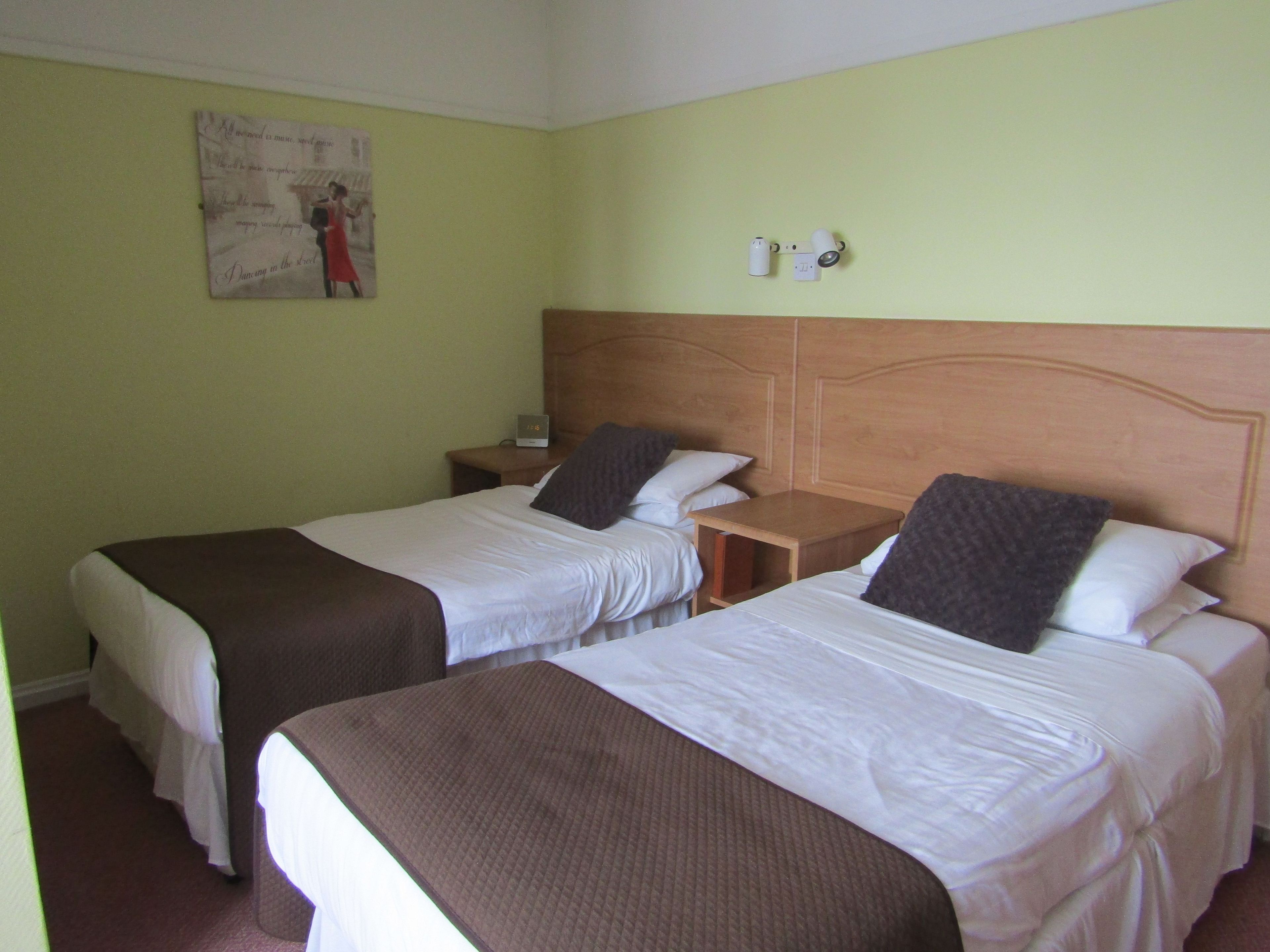 A hotel room with two single beds, a wooden headboard, a nightstand with a lamp, and a wall clock.