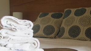 Une pile de serviettes blanches, soigneusement pliées, est posée sur un lit dont la tête de lit est marron et qui est agrémenté d’oreillers à motifs.