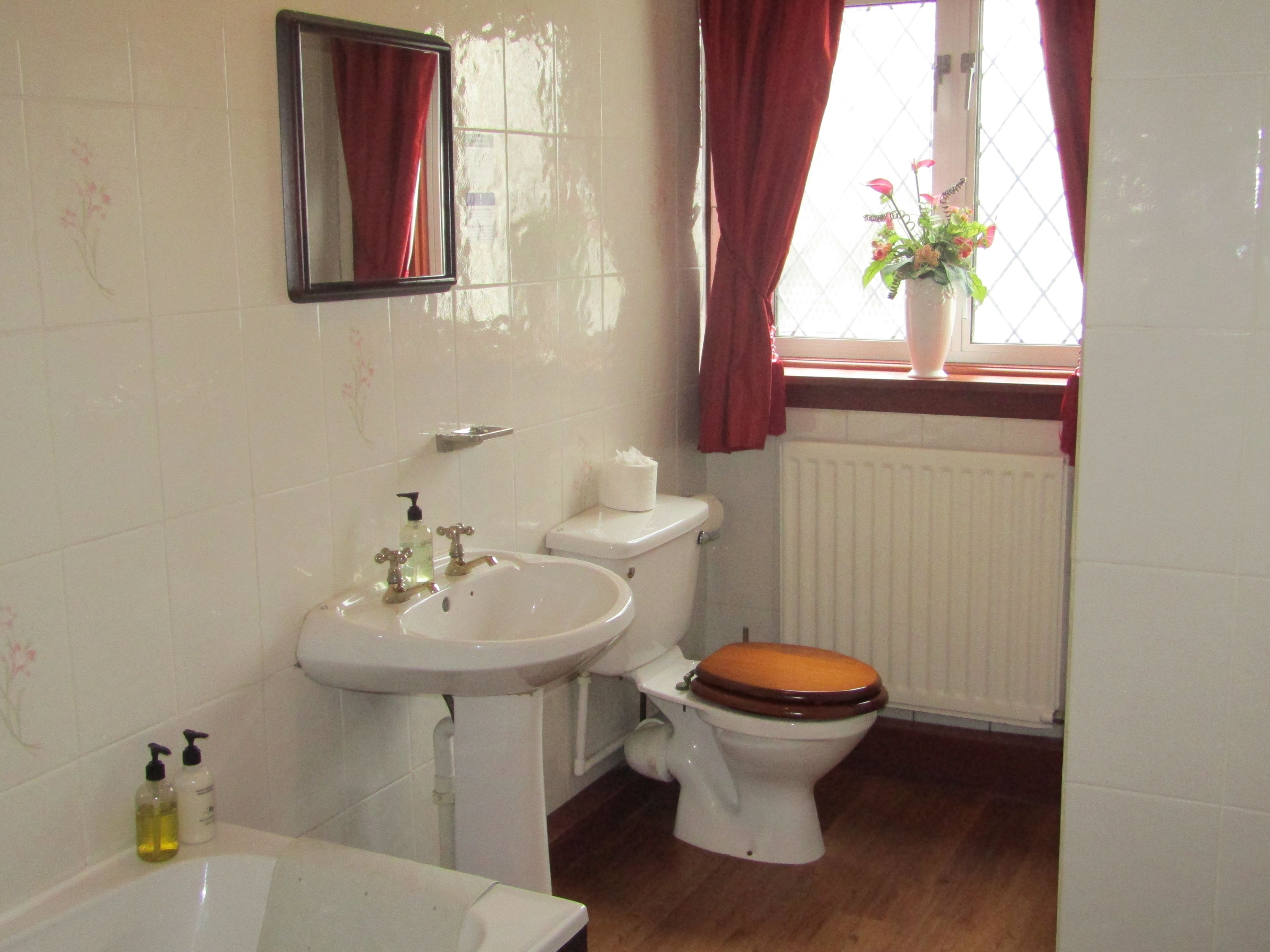 A bathroom with a white sink, a mirror, a toilet with a wooden seat, and a window with red curtains.