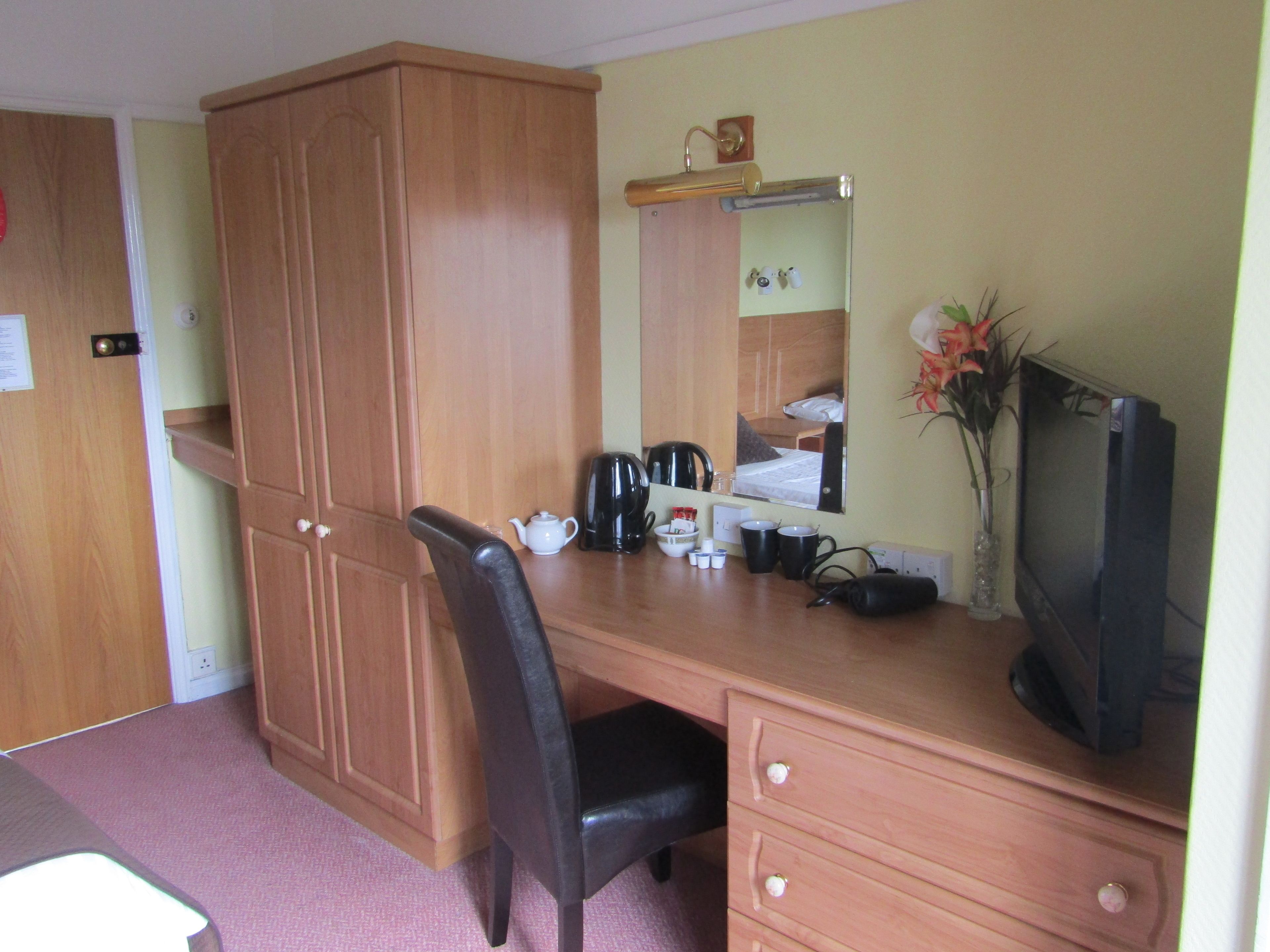 A hotel room with a wooden desk, a black chair, a television, a mirror, and a small cabinet.