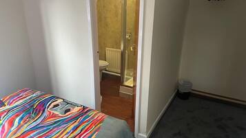 A bedroom with a colorful bedspread, a visible bathroom with a shower, and a small trash bin.