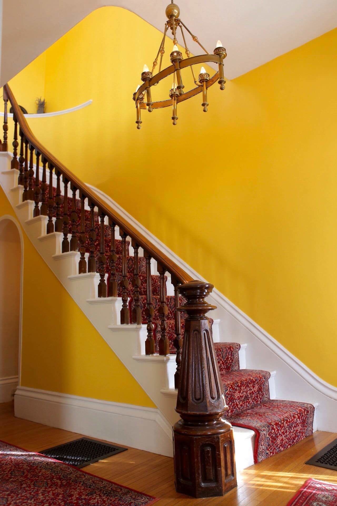 Una escalera con barandales blancos y balaustres de madera, que conduce a una habitación con paredes amarillas y un candelabro.