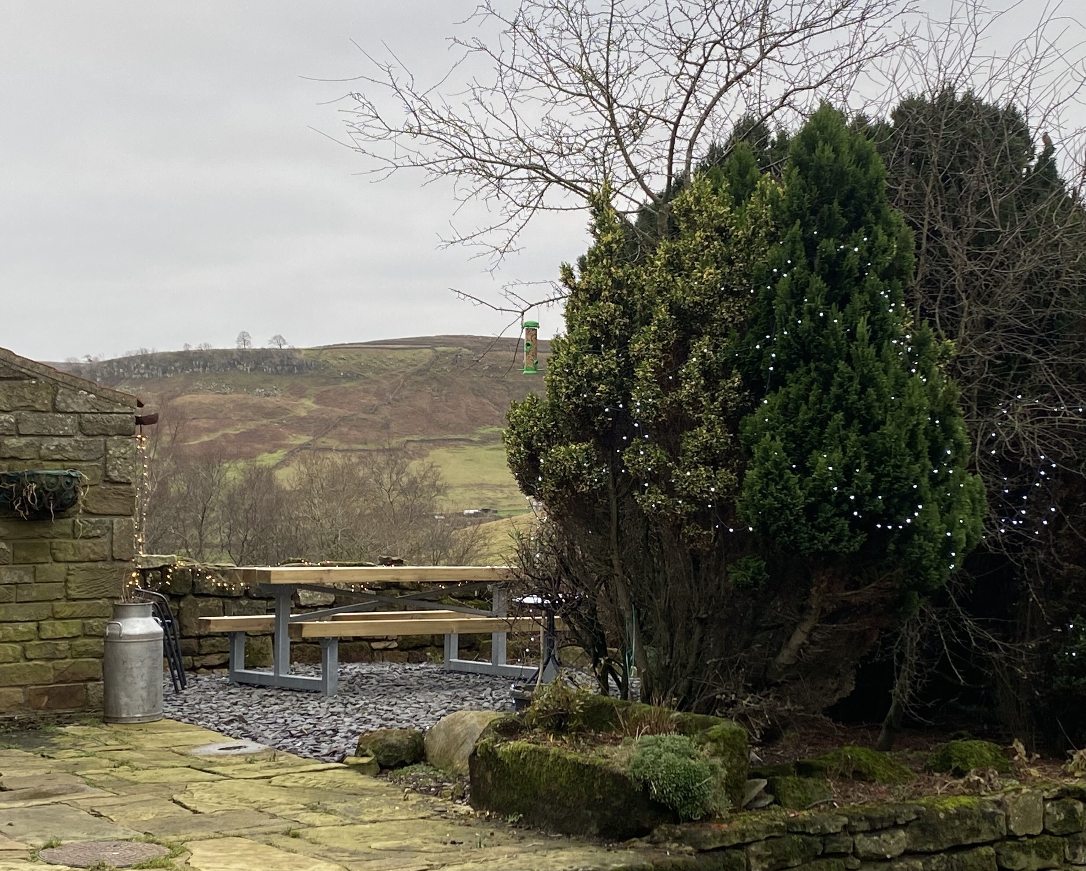 A stone building with a bench and a tree in the foreground, a stone wall, and a view of a hill with a wind turbine in the distance.