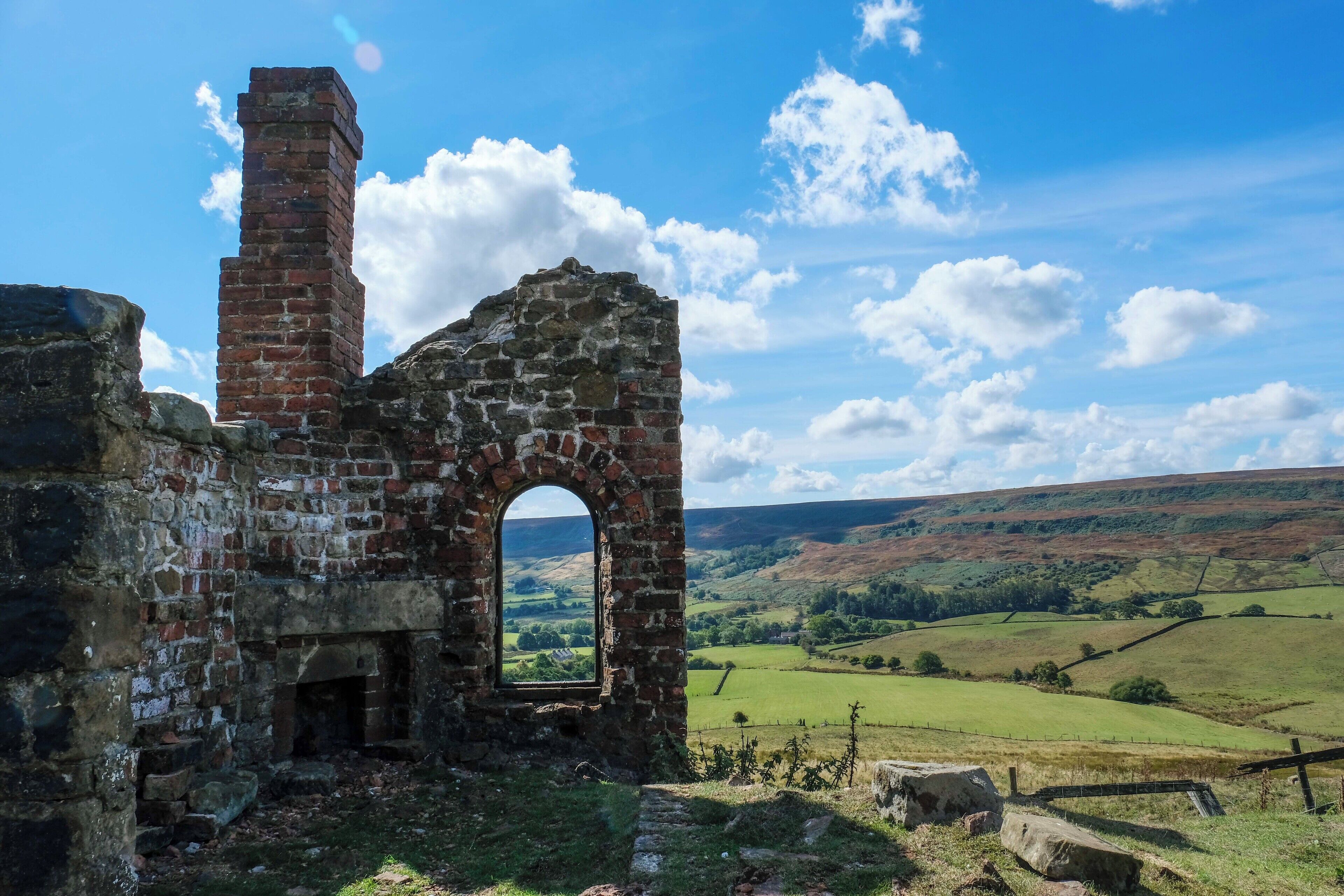 An old stone structure with a brick chimney and an arched window, surrounded by a rural landscape with rolling hills and a clear sky.