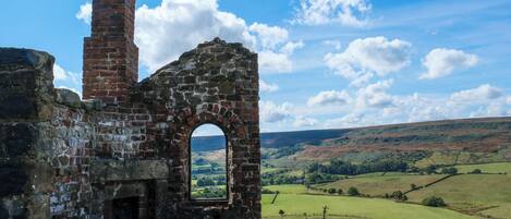 An old stone structure with a brick chimney and an arched window, surrounded by a rural landscape with rolling hills and a clear sky.