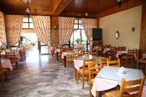 A restaurant with wooden tables and chairs, large windows with curtains, and a tiled floor. - Poseidon Hotel (Malevizi)