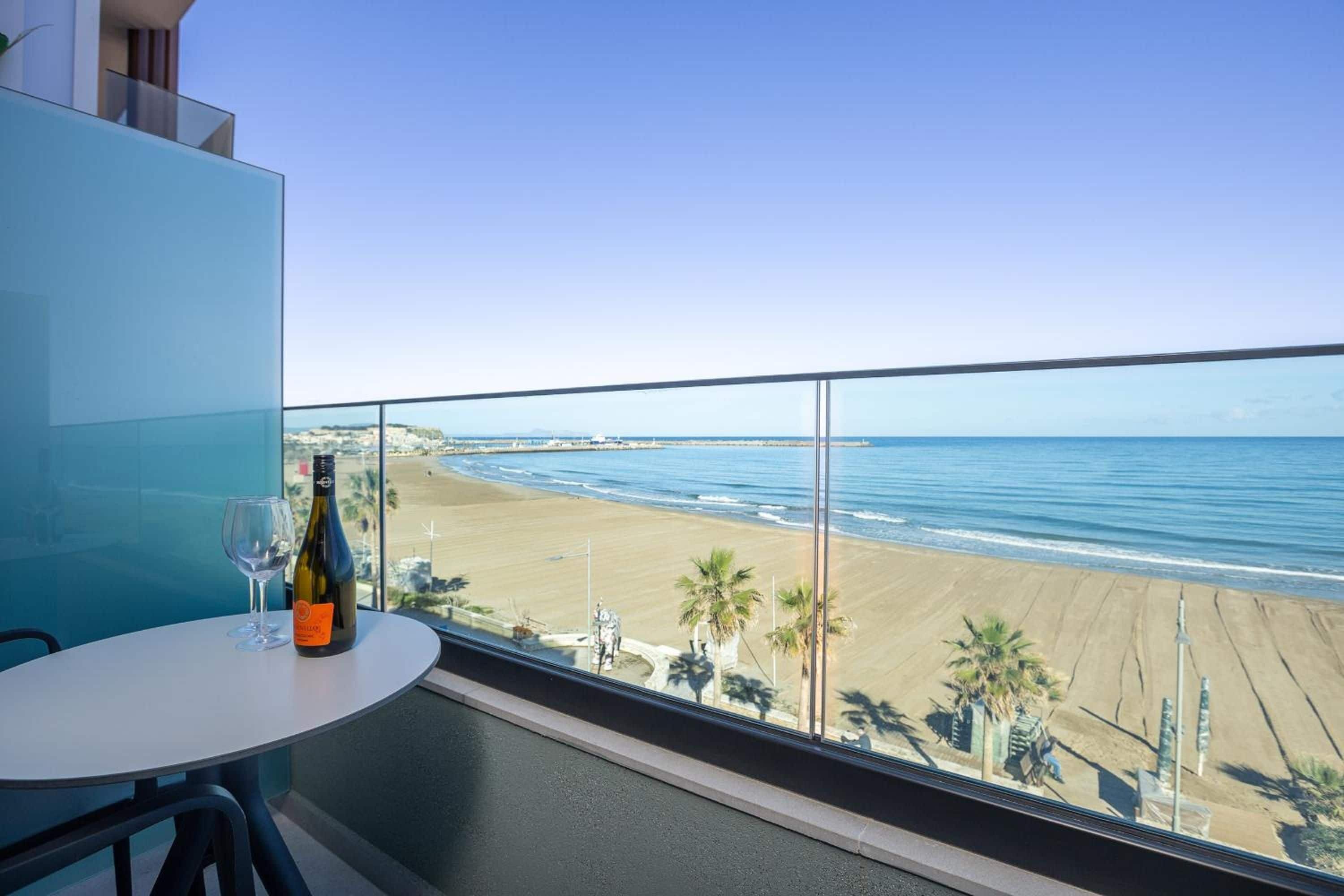 A balcony with a table, a bottle of wine, and a glass. The view includes a beach and the ocean.