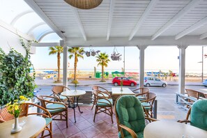 An outdoor seating area with wicker chairs and tables, overlooking a street with cars and palm trees. - Poseidon Hotel (Malevizi)