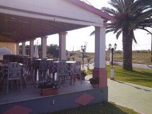 An outdoor patio with tables and chairs, a covered area, and a palm tree.