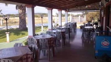 Un patio exterior con mesas y sillas, un carrito azul con un logotipo y vistas al paisaje.