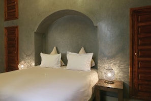 A bedroom with a bed, two pillows, a nightstand with a lamp, and wooden doors. - Riad L'emir (Marrakech)