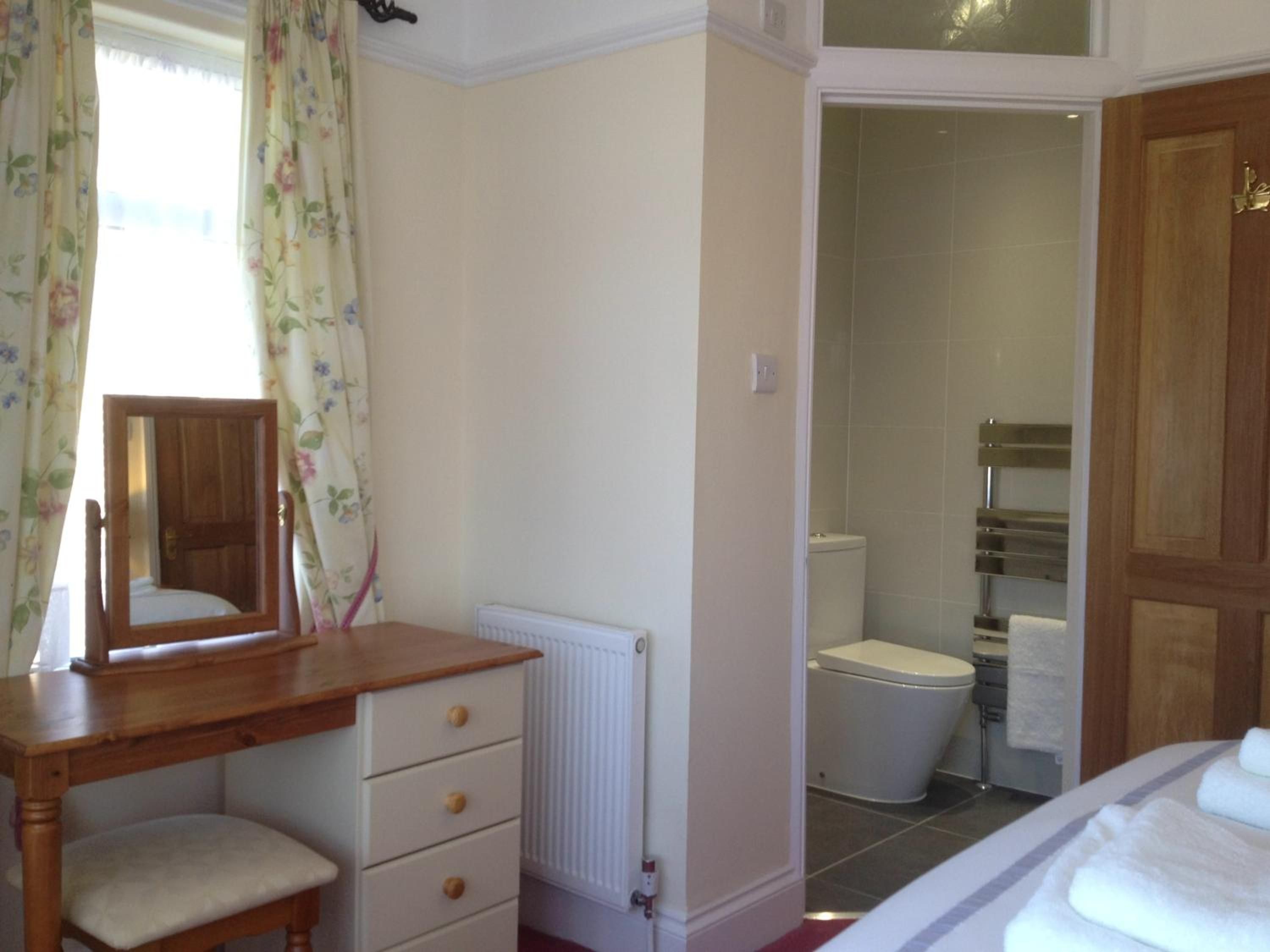 Une chambre avec une commode en bois, un miroir, un radiateur, un lit avec une literie blanche, et une salle de bain avec des toilettes et un porte-serviettes.