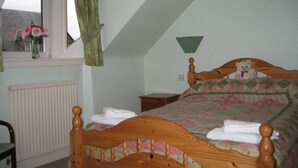 A bedroom with a sloped ceiling, a wooden bed with a headboard, a bedside table, a nightstand, a window with curtains, and a radiator. - Mardon Guest House (Inverness)