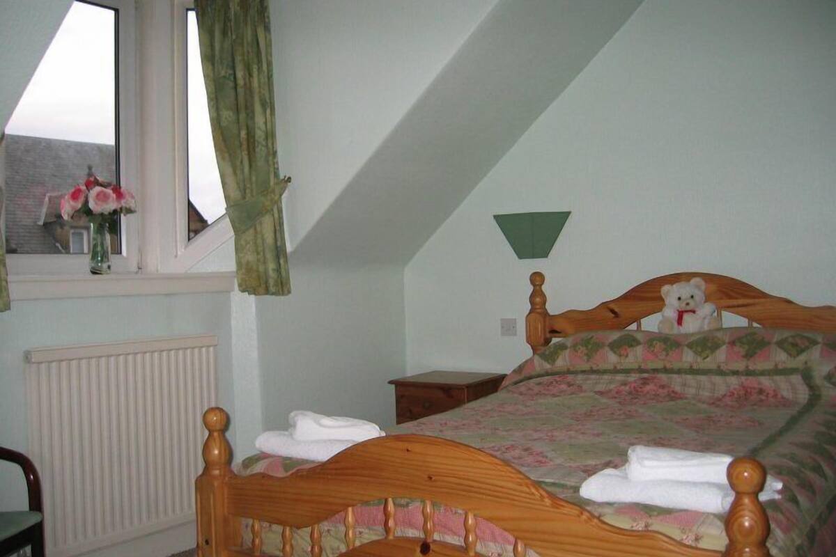 A bedroom with a sloped ceiling, a wooden bed with a headboard, a bedside table, a nightstand, a window with curtains, and a radiator.