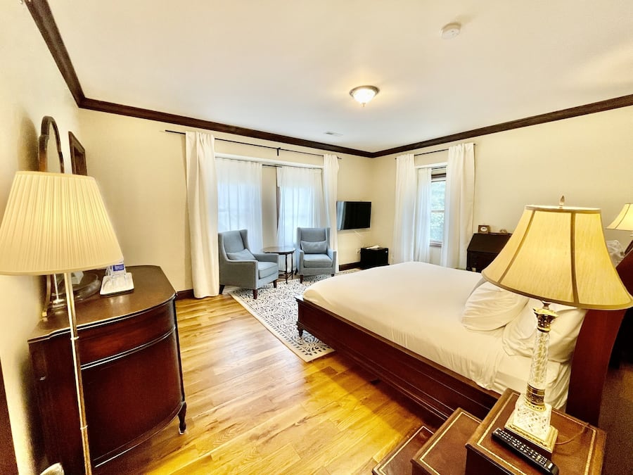 Quarto de hotel com uma cama grande, duas luminárias de cabeceira, uma escrivaninha com abajur, uma cadeira, uma televisão e uma janela com cortinas.