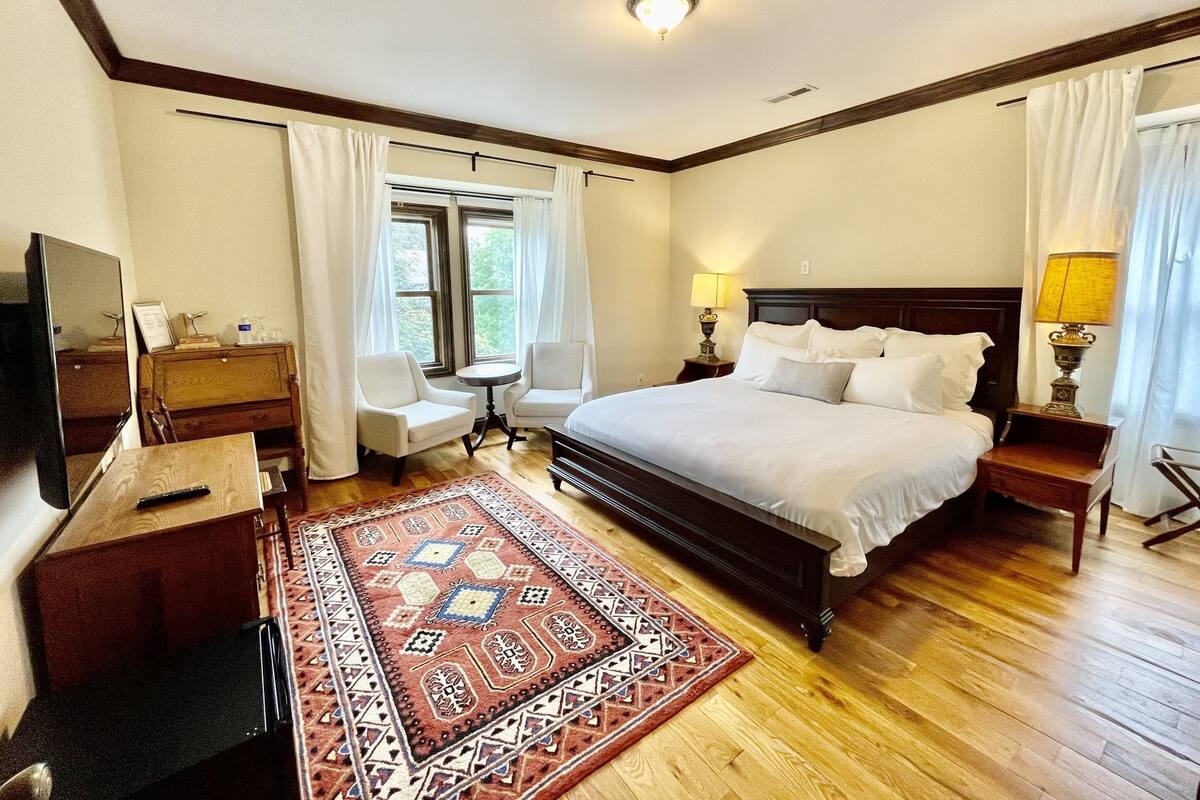 Ein Schlafzimmer mit einem großen Bett, zwei Nachttischen, einem kleinen runden Tisch, einem Sessel, einer Kommode, einem Fernseher und einem Fenster mit Vorhängen.