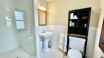 A bathroom with a glass shower enclosure, a pedestal sink, a toilet, and a vanity with shelves.