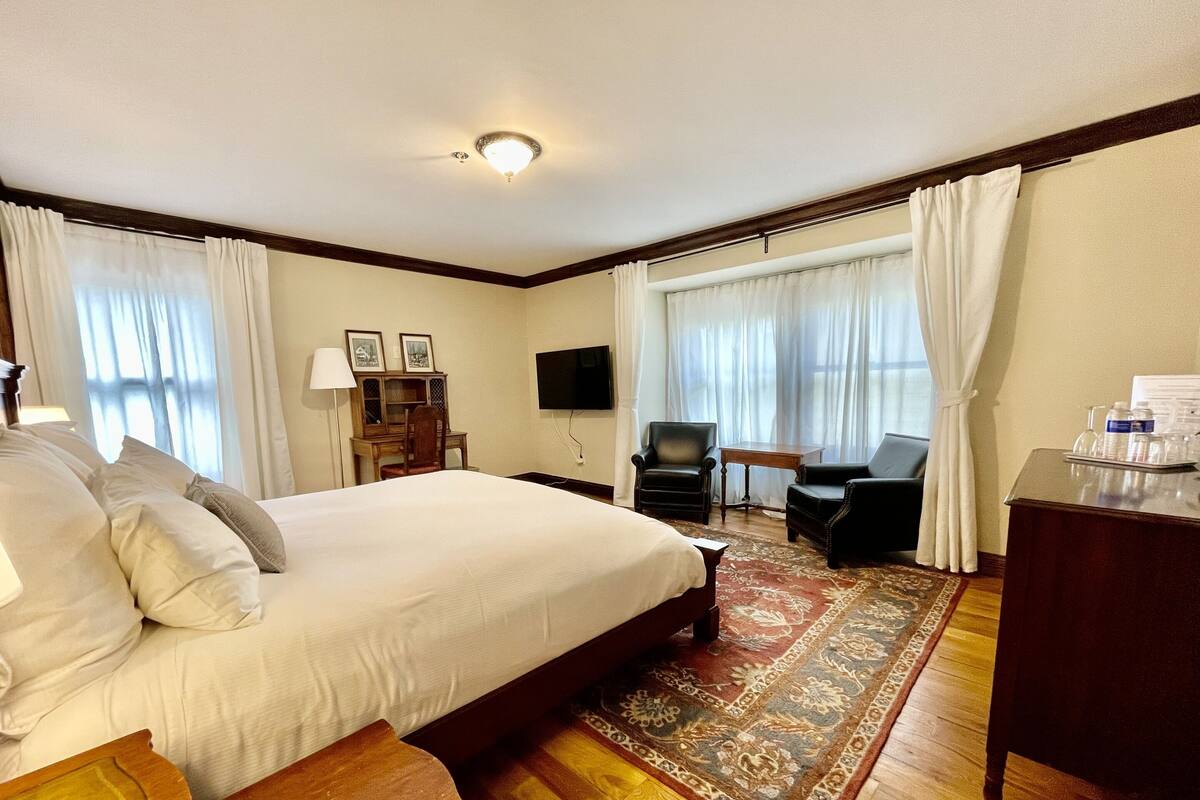 Ein Hotelzimmer mit einem großen Bett, einem Schreibtisch, einem Sessel, einem Fernseher und einem Schreibtisch mit einer Wasserflasche.