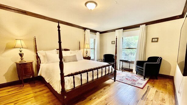 Un dormitorio con una cama con dosel, una mesita de noche con lámpara, una silla y una ventana con cortinas.