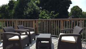 Una terraza de madera con mobiliario de mimbre, incluyendo sillas y una mesa, rodeada de árboles.