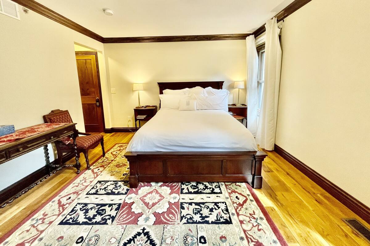 Ein Hotelzimmer mit einem großen Bett, einem hölzernen Kopfteil, zwei Nachttischen mit Lampen, einem Schreibtisch aus Holz und einem gemusterten Teppich.