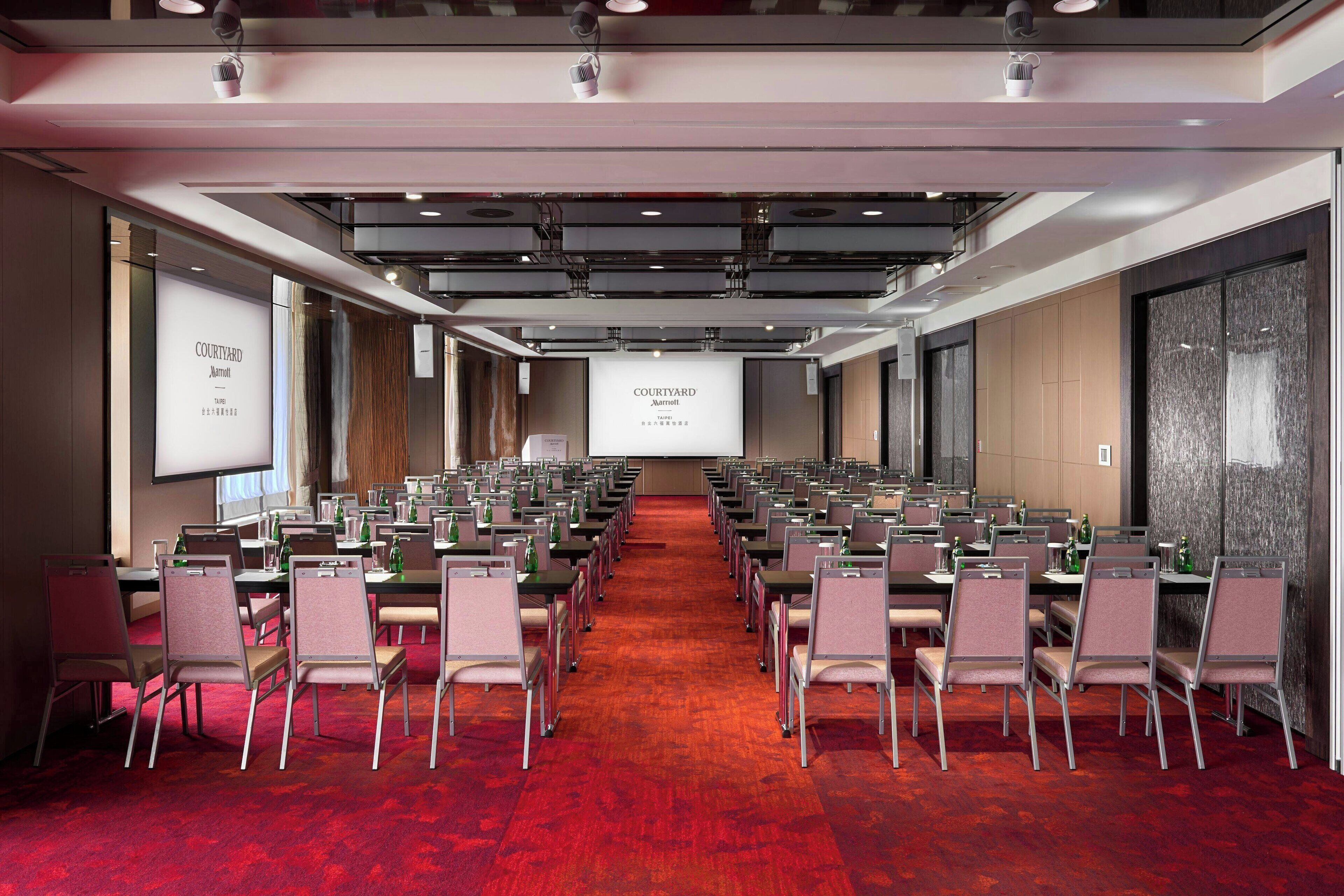 Un salón de conferencias con filas de mesas y sillas, una pantalla grande al frente y un piso cubierto de alfombra roja.