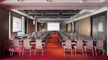 Un salón de conferencias con filas de mesas y sillas, una pantalla grande al frente y un piso cubierto de alfombra roja.