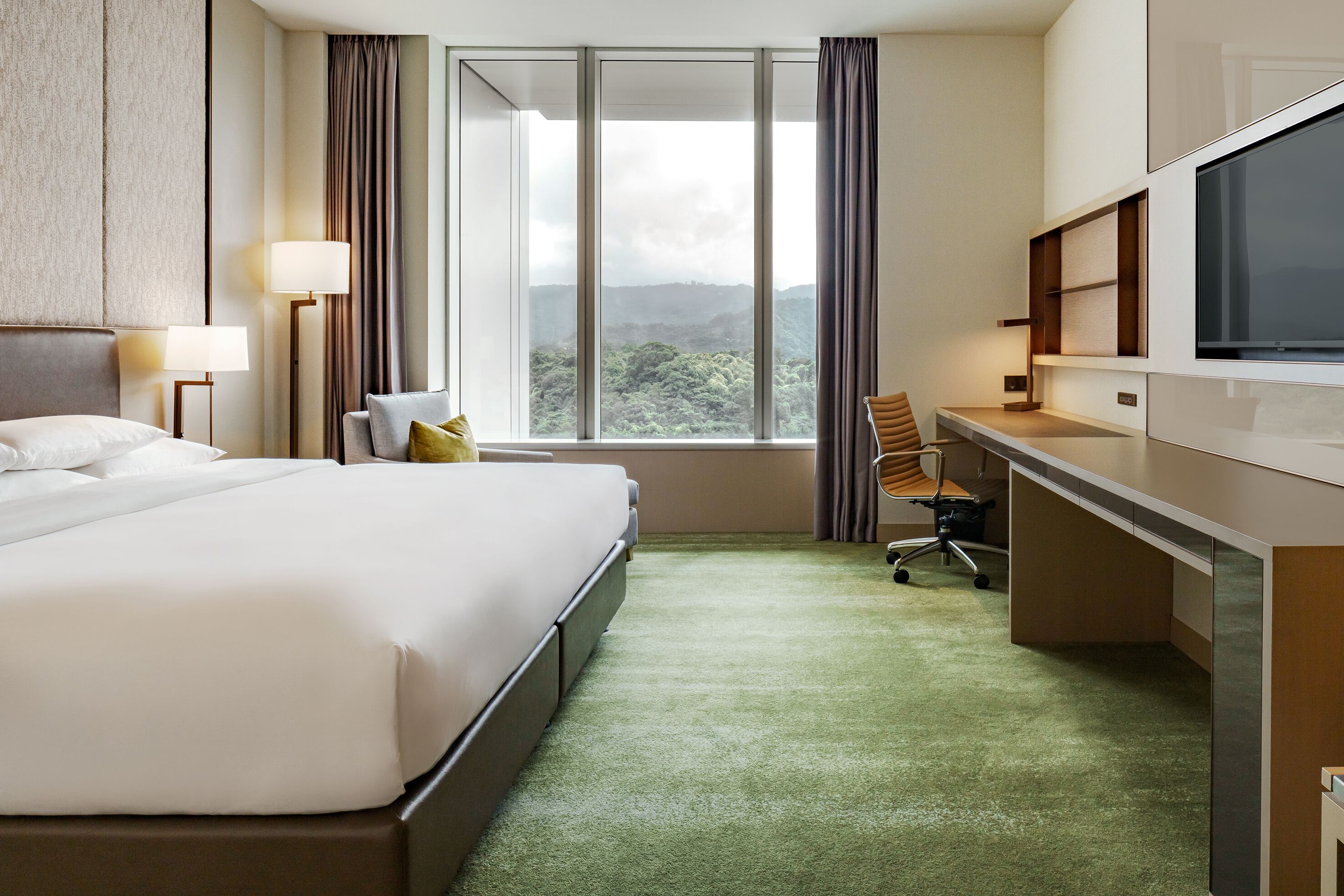 A hotel room with a large bed, a desk, a chair, a TV, and a view of the mountains through the window.