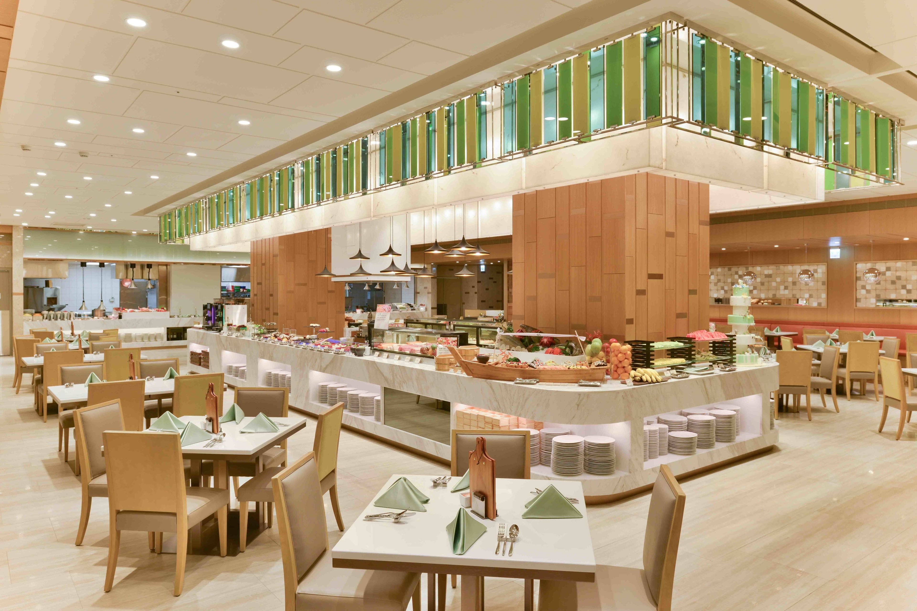 Uma ampla sala de jantar com várias mesas e cadeiras, um balcão de buffet com diversos alimentos, e teto alto com iluminação embutida.