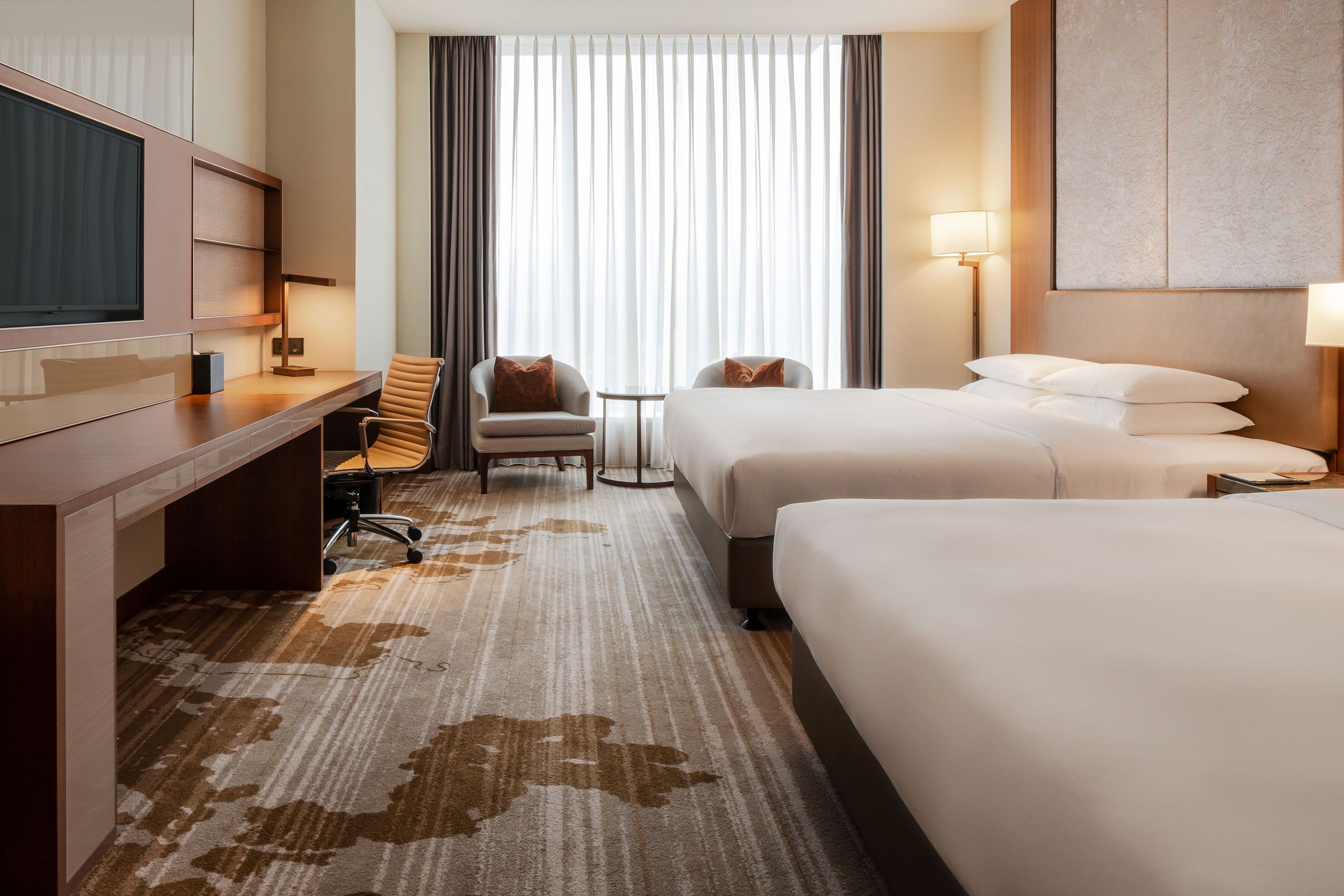 A hotel room with two beds, a desk, a chair, a TV, and a large window with curtains.