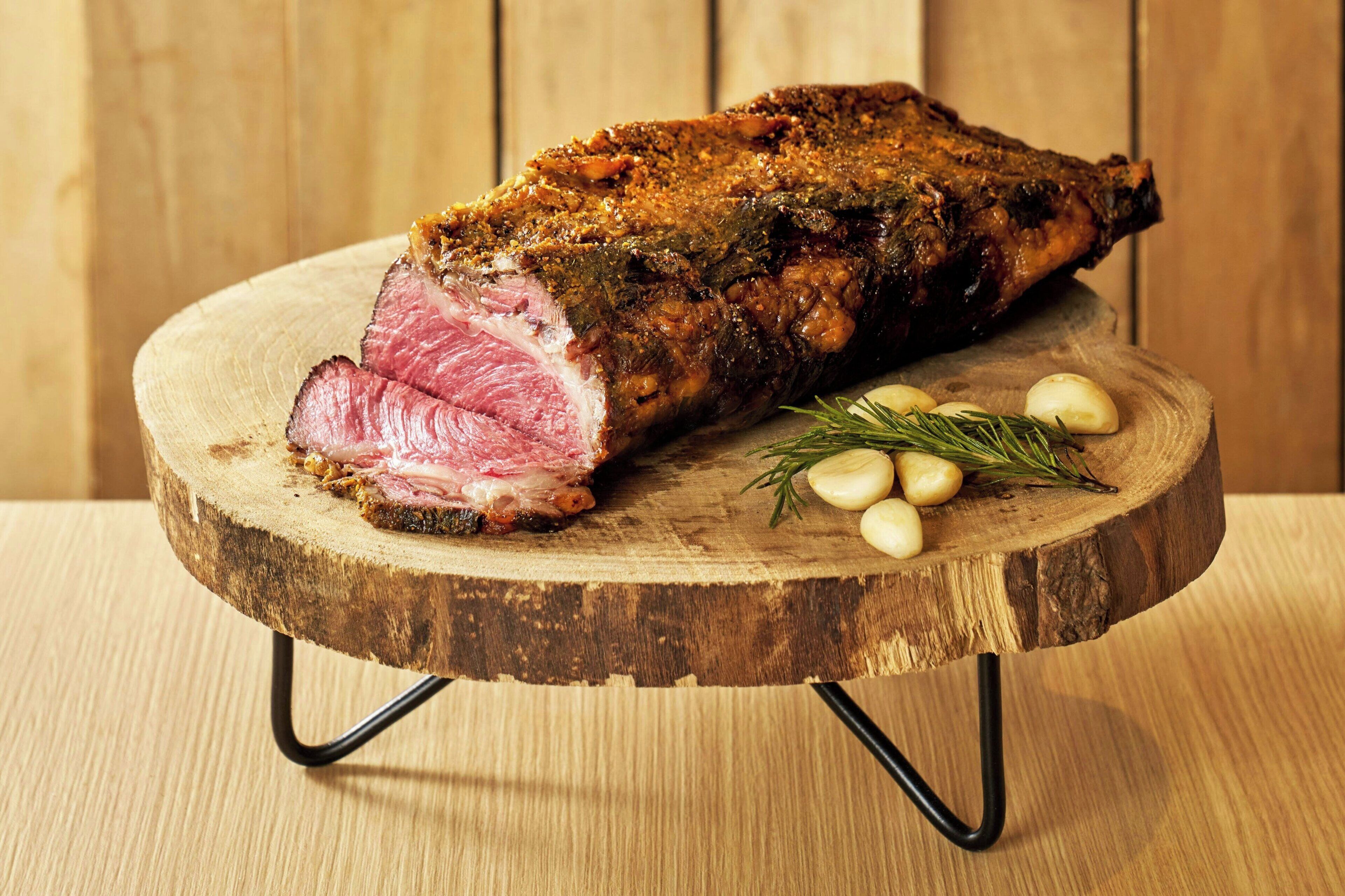 A large piece of meat on a wooden cutting board with garlic cloves and rosemary.