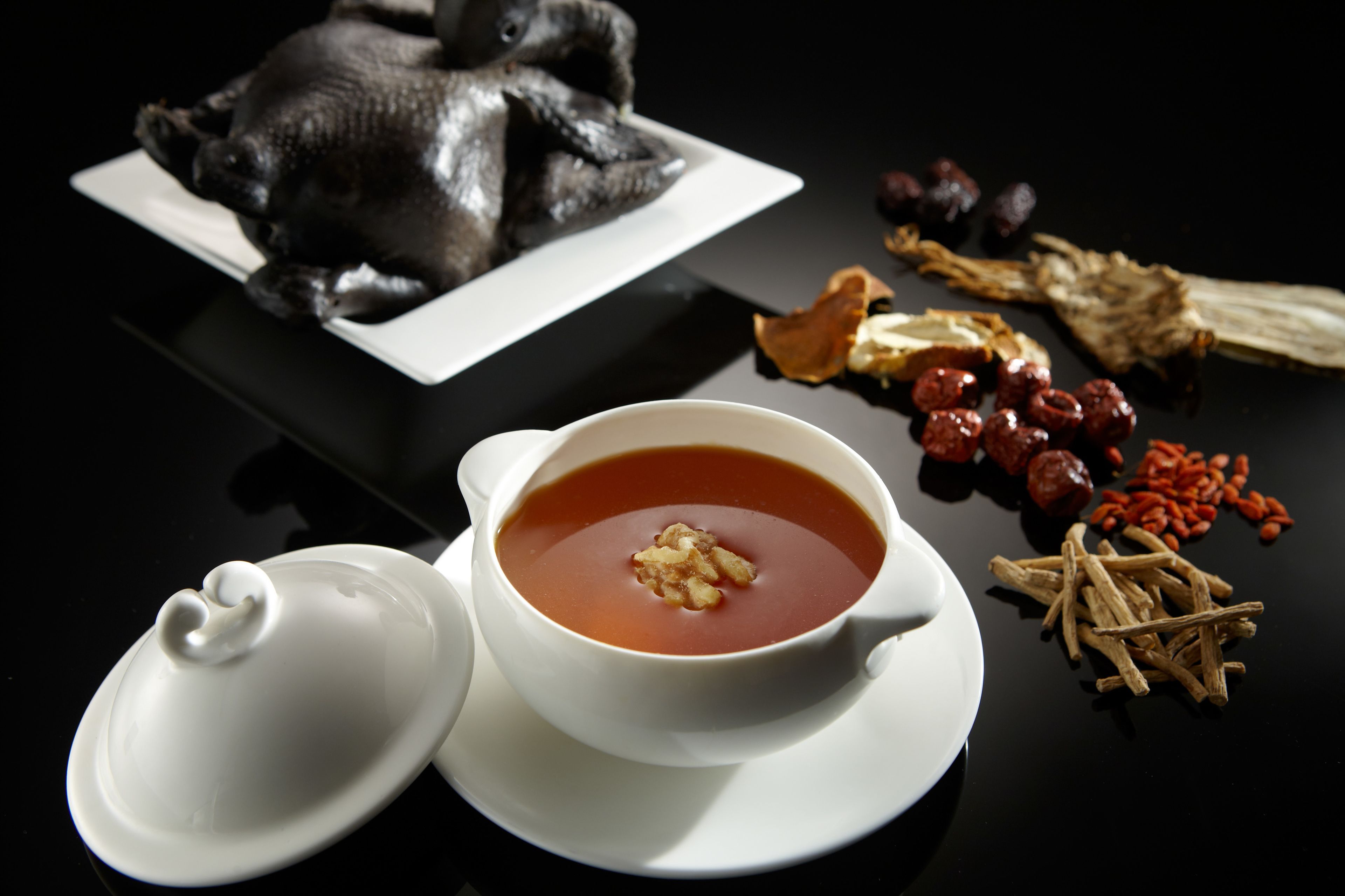 A white teapot with a lid, a white teacup with a saucer, a bowl of red soup with a garnish, and various dried ingredients on a black surface.