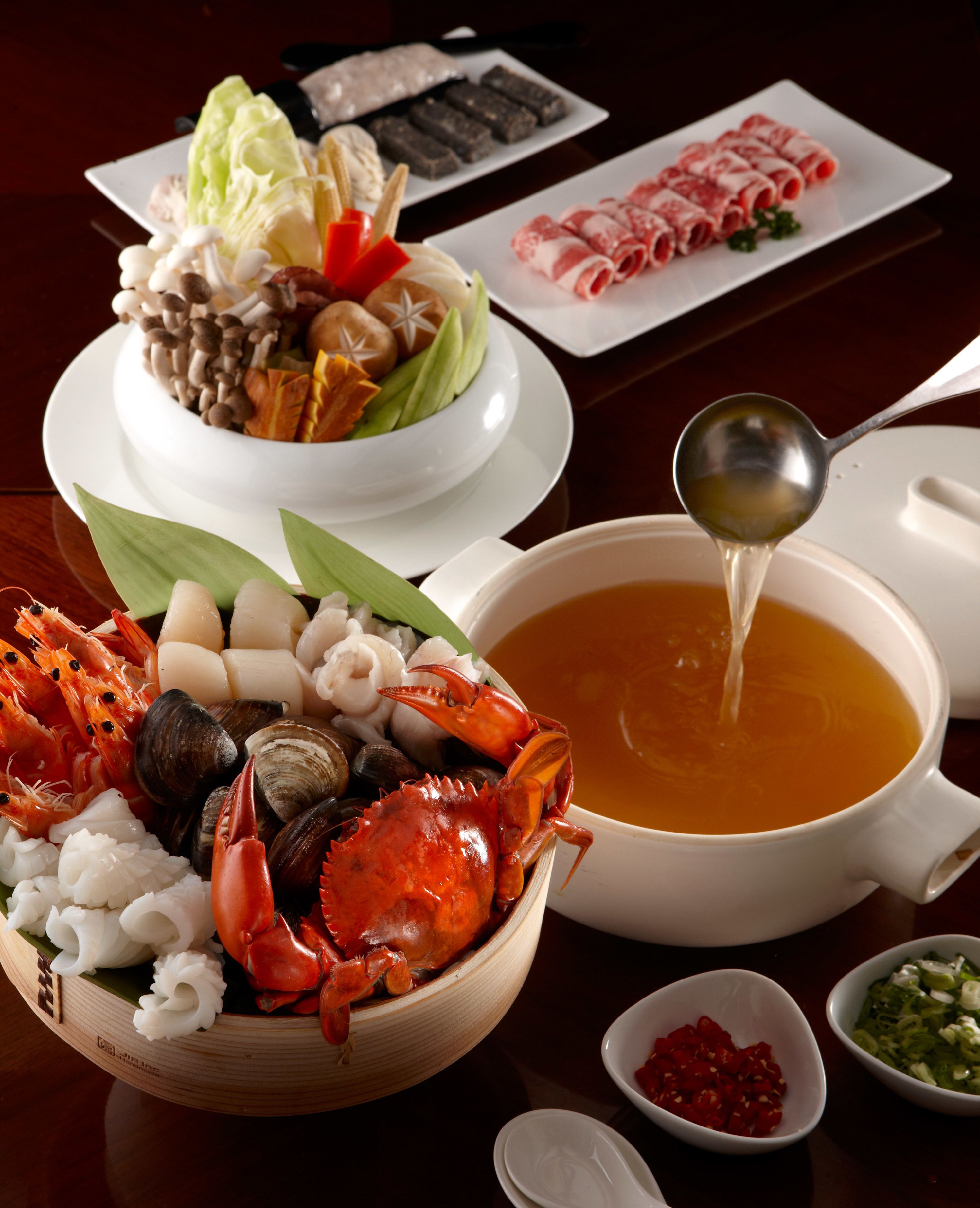 A table with a variety of seafood, including a crab, clams, and shrimp, alongside a bowl of soup and a plate of sliced meat.