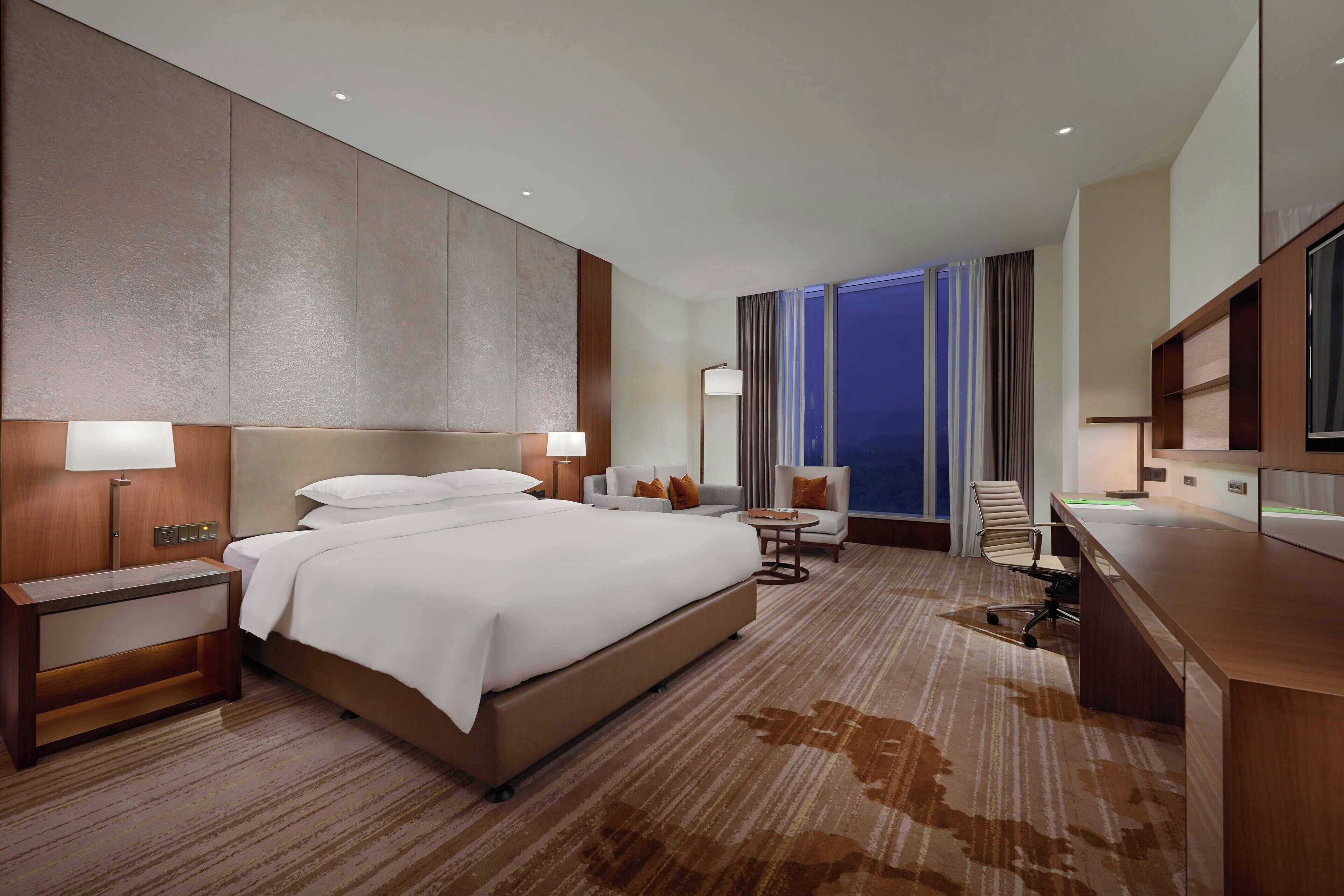 A modern hotel room with a large bed, a desk, a chair, and a view of the city at night.